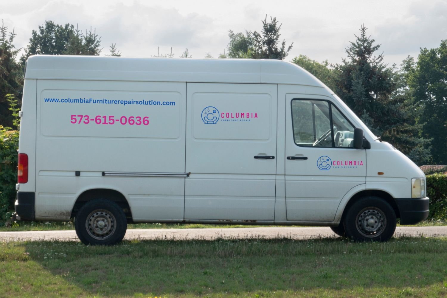 A white delivery van displaying branding and contact details, parked in a grassy field with trees and a cloudy sky behind it.