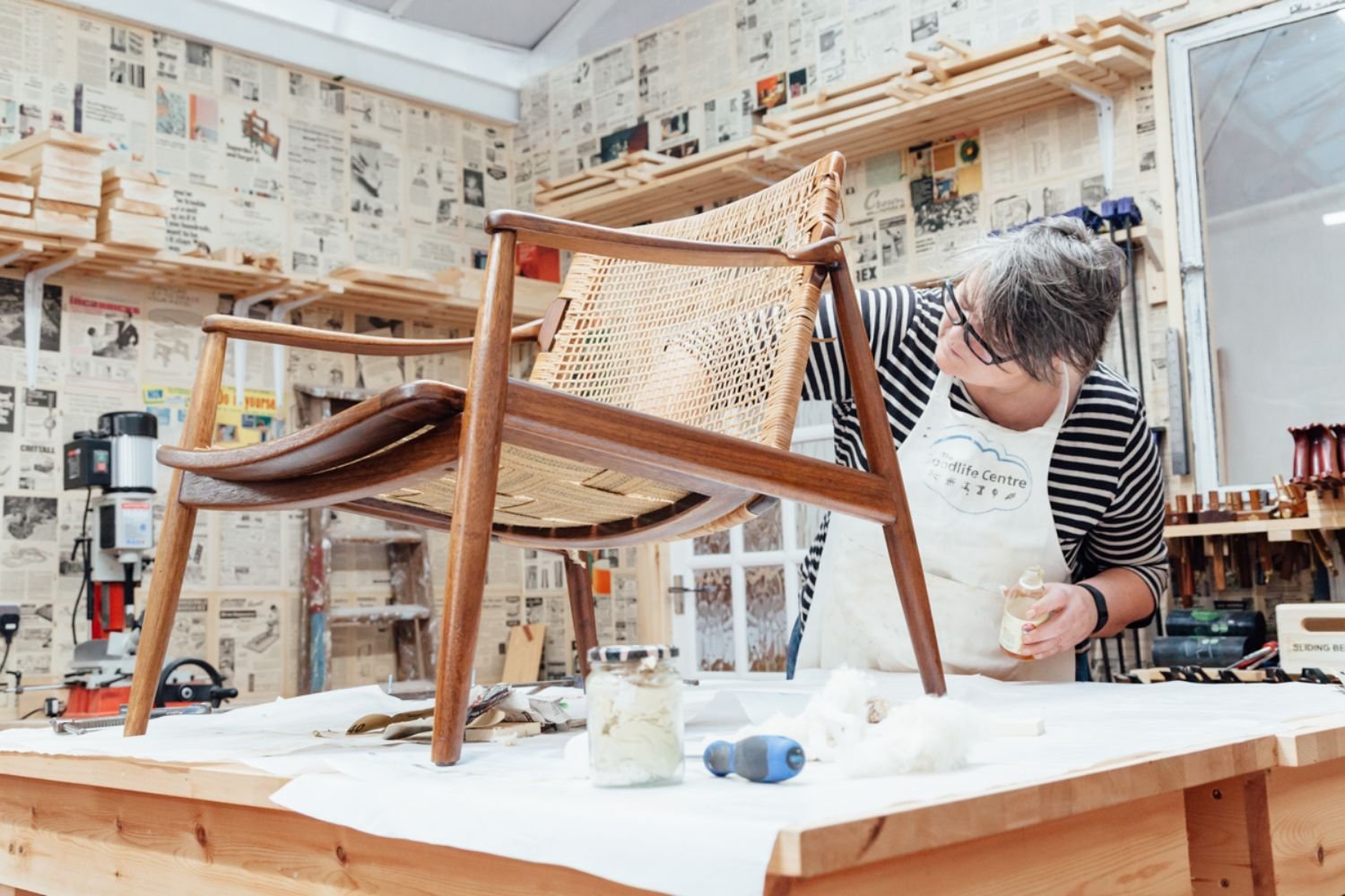 A woman works on refurbishing a wooden chair in a busy workshop, surrounded by an array of tools and materials.