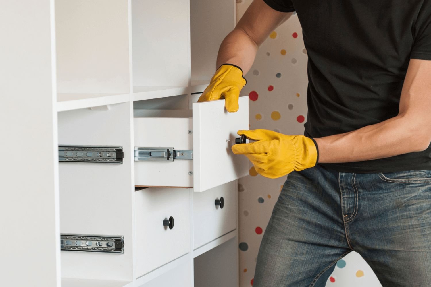 A man in a black shirt and yellow gloves installs a drawer into a white cabinet.