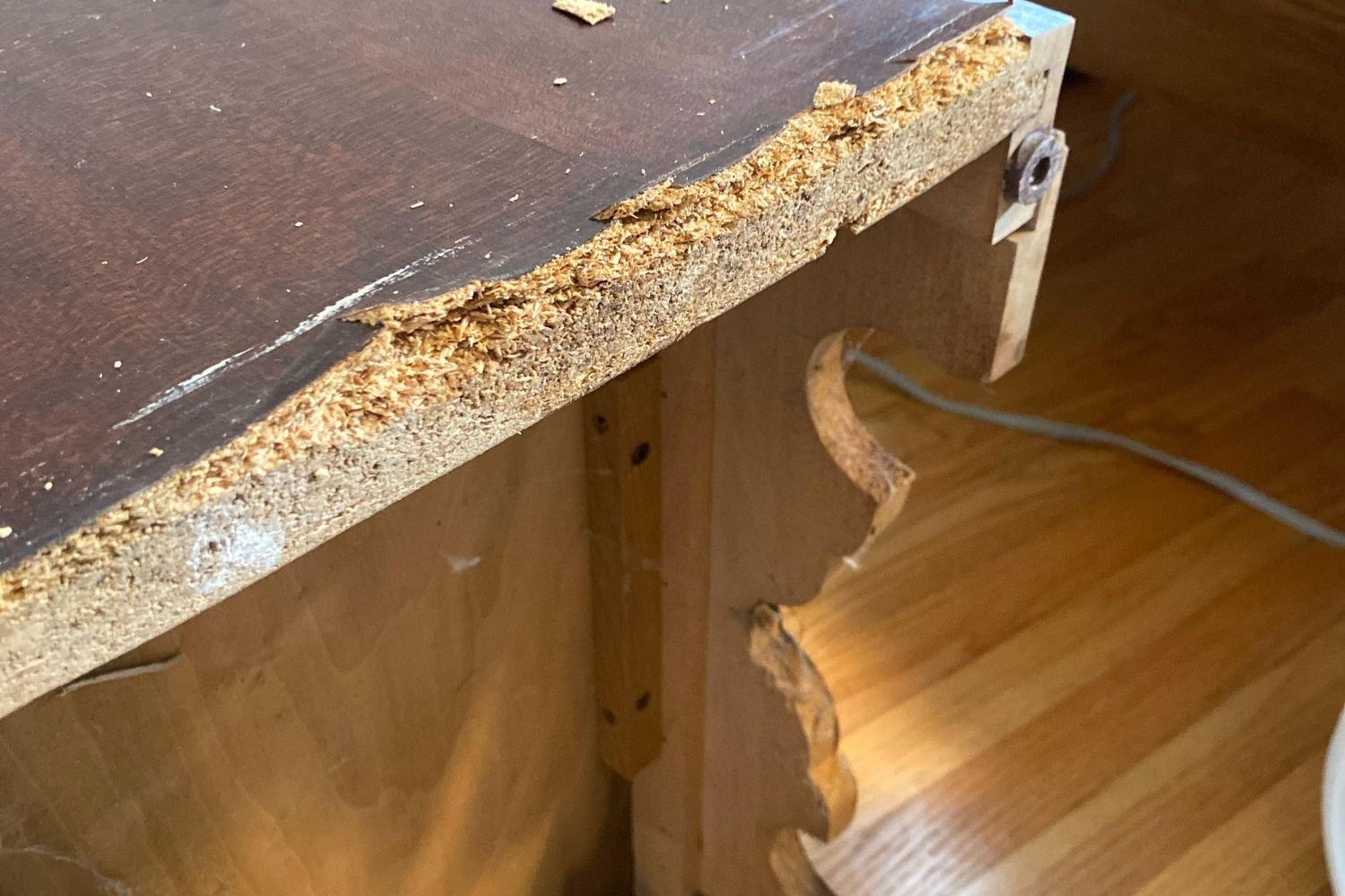A damaged wooden desk edge with chips and splinters, revealing a wooden floor and a visible power cable underneath.