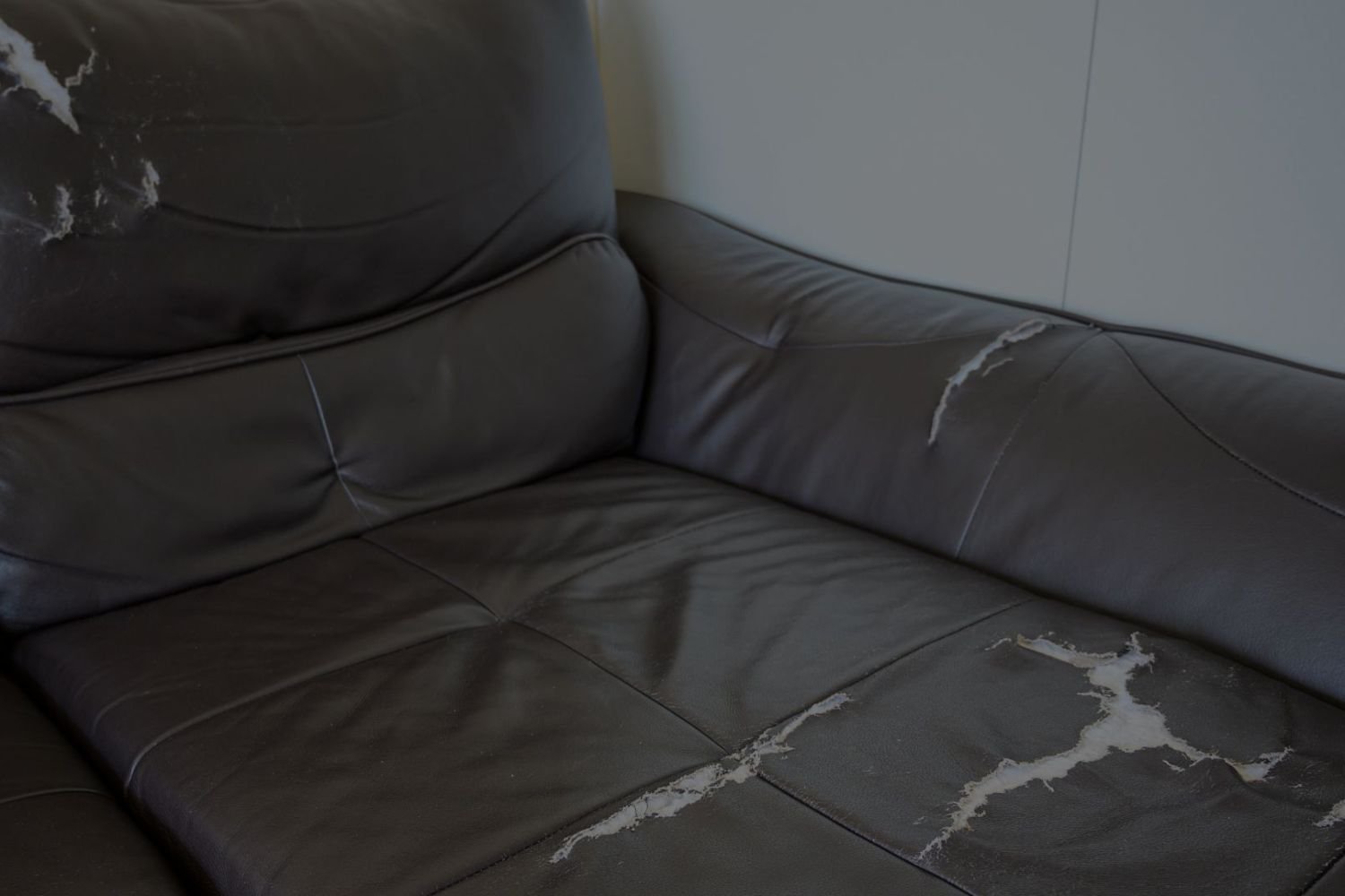 A worn gray leather couch with visible cracks and a white stain, situated in a modestly lit space.