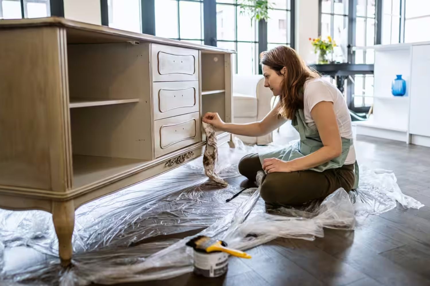 A woman focused on painting a dresser in a bright room, with paintbrushes and color swatches nearby.